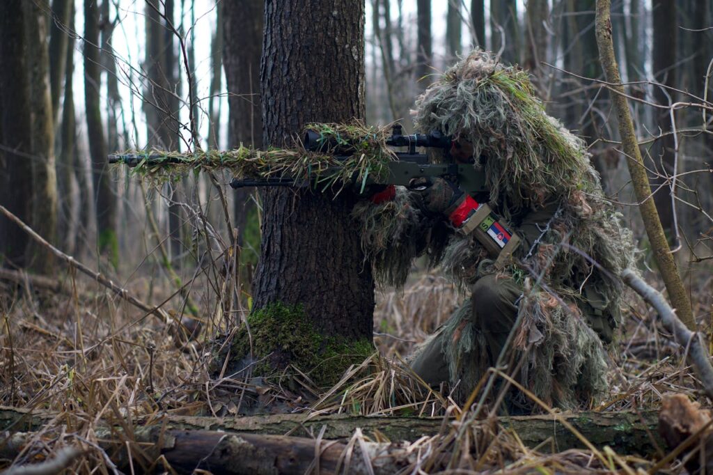 Hunter in a ghillie suit sitting beside a tree and preparing to take a shot.