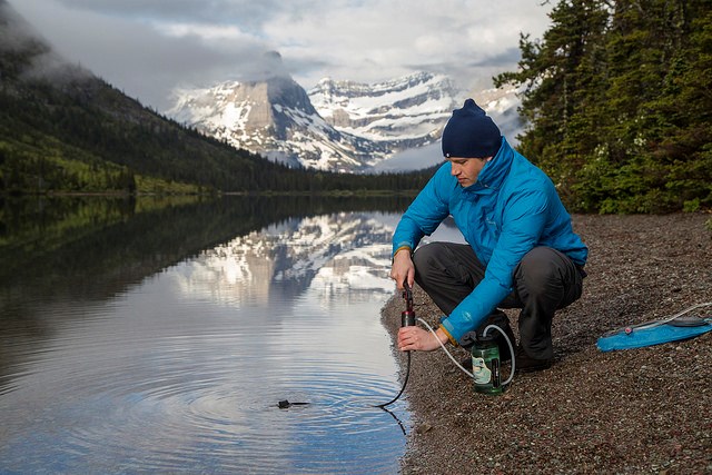 a man kneeling next to a mountain lake and filtering drinking water